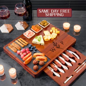 May include: A wooden serving board with a sliding drawer that holds a set of cheese knives. The board has a slate insert and is engraved with the initials "W.P.". The board is filled with various snacks, including cheese, grapes, crackers, and olives.