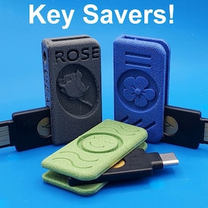 May include: Three 3D-printed plastic USB drive covers in gray, blue, and green. The gray cover features the text ROSE and a rose graphic. The blue cover has a flower design, and the green cover has a smiley face. The text Key Savers! is at the top.