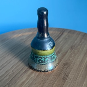May include: A small, ceramic bell with a dark handle and a flared base. The bell features a color scheme of black, green, and brown. The bell is sitting on a wooden surface with a blue background.