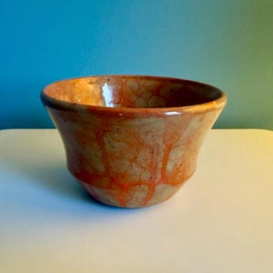 May include: A handmade ceramic bowl with a wide, flared shape. The bowl features a speckled, tan base color with vibrant orange drips and accents. The interior is a glossy, warm brown. The bowl is approximately 6 inches in diameter and 4 inches tall.
