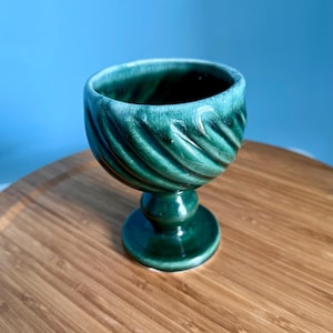 May include: A green ceramic goblet with a spiral design. The goblet has a rounded bowl shape and sits on a pedestal base. The interior is dark, and the exterior has a glossy finish. The goblet is on a wooden surface.