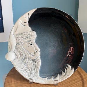 May include: A round ceramic plate featuring a raised, white relief sculpture of a bearded Santa Claus profile on the left side. The remainder of the plate has a smooth, deep blue-black glazed finish that transitions into the white sculpted beard.