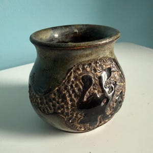 May include: A small, round ceramic pot with a wide, flared rim. The exterior features a mottled, dark green and brown glaze with a textured, rough surface pattern that includes a smooth, dark brown, raised abstract shape on the front side.