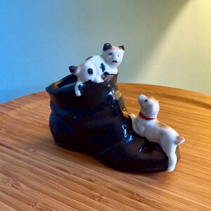 May include: A black ceramic boot planter with two small dog figurines. One dog is white with black spots, and the other is white with brown spots and a red collar. The boot is on a wooden surface.