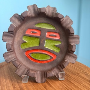 May include: A brown ceramic ashtray with a stylized face design. The face is made of green and red glaze. The ashtray has a scalloped edge.