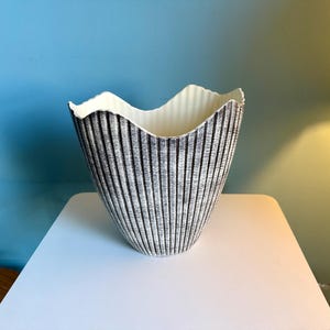 May include: A decorative ceramic vase with a textured, vertically striped exterior in shades of gray and black. The rim has a wavy, scalloped edge and a white interior. The vase is set on a white surface.