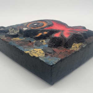 May include: A colorful ceramic wall sculpture with a red, orange, and black abstract design. The sculpture is mounted on a black wooden base.