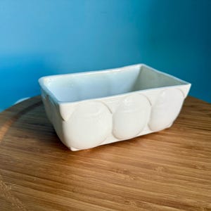 May include: A rectangular, off-white ceramic container with a rounded design on the front. The container is set on a wooden surface against a blue background. The container is likely used for storage or display.