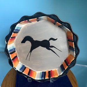 May include: A decorative ceramic plate with a black scalloped edge and a central image of a black horse. The plate's rim features a colorful striped pattern in orange, blue, and white. The plate is displayed on a wooden surface.