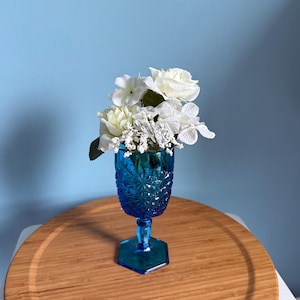 May include: A vibrant blue glass goblet with a textured pattern, filled with white flowers. The goblet has a hexagonal base and a slender stem. The flowers are a mix of white roses and smaller white blossoms, creating a striking contrast.