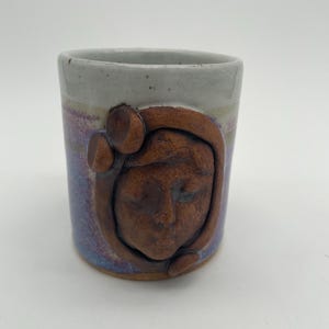May include: A ceramic mug with a brown face sculpted on the side. The mug has a white rim and a purple and white glaze.