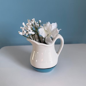May include: A small white ceramic pitcher with a blue base, holding white flowers. The pitcher has a handle and a curved spout. The flowers include small white buds and larger white petals.