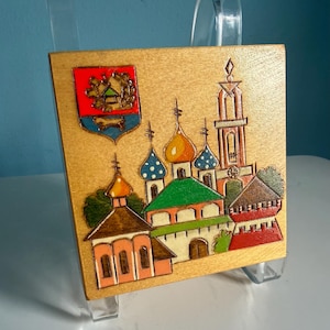 May include: A square wooden plaque featuring a pyrography and painted illustration of a cathedral with multiple domes and a tall bell tower. A colorful coat of arms with red, blue, and gold details is positioned in the upper left corner.