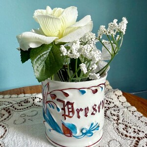 May include: A white ceramic mug with the word "Bresm" in burgundy lettering, adorned with blue and burgundy floral designs. The mug holds white flowers and greenery, creating a decorative display. The mug is on a white lace doily.