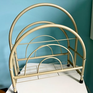 May include: A gold-colored metal magazine rack with a curved design. The rack has multiple sections for organizing reading materials. The frame is made of thin metal rods and has a sturdy base with black feet.