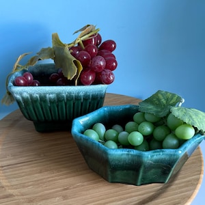 May include: Two teal ceramic bowls filled with grapes sit on a wooden surface. One bowl holds red grapes, the other green. The bowls have a textured, ribbed design and are set against a blue background.
