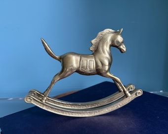May include: A silver-colored rocking horse figurine. The horse is in a galloping pose, with its legs extended. The rocking base has a curved design. The figurine is made of metal and has a detailed mane and tail.
