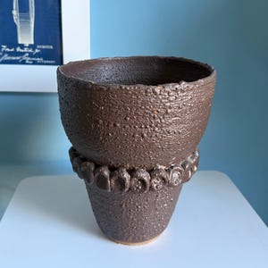 May include: A dark brown ceramic vessel with a rough, textured surface and a wide, open top. The exterior features a decorative band of small, raised circular clay elements encircling the middle section of the pot, creating a distinct visual contrast.