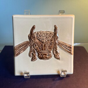 May include: Square ceramic tile with a raised relief of a bull's head. The bull's head is brown with detailed horns and a textured face, set against a cream-colored background. The tile is displayed on a clear stand.