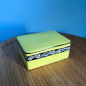 May include: A square, yellow trinket box with a black and white decorative border. The box is sitting on a wooden surface against a blue background. The box has a matching yellow lid.