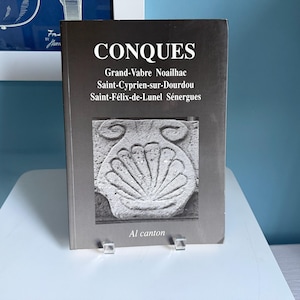 May include: A softcover book with a dark gray cover featuring the white text CONQUES, Grand-Vabre Noailhac, Saint-Cyprien-sur-Dourdou, Saint-Félix-de-Lunel Sénergues, and Al canton. A black and white photograph of a stone scallop shell carving.