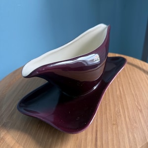 May include: A deep plum-colored gravy boat with a smooth, glossy finish and a contrasting cream-colored interior. The vessel features a curved, elongated spout and sits securely on a matching rectangular underplate with rounded edges.