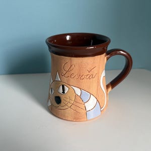May include: A ceramic mug with a brown rim and handle, featuring a tan body with a cartoon cat design. The cat has blue and white stripes, and the word "Levoca" is written on the mug.