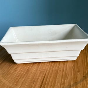 May include: A rectangular, off-white ceramic planter with a stepped design. The planter is empty and sits on a round, wooden cutting board. The background is a solid blue.