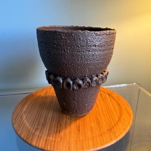 May include: A dark brown ceramic planter with a textured surface and a band of raised circles around the middle.