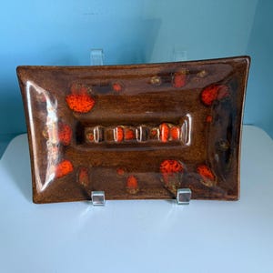 May include: A rectangular ashtray with a brown base and vibrant orange accents. The ashtray has a glossy finish and a recessed area with multiple rests for cigarettes. The design is reminiscent of mid-century modern decor.