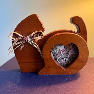 May include: Wooden cat-shaped photo holder with a heart-shaped opening. The holder is brown with a decorative bow and a floral fabric visible through the heart. A unique home decor item.