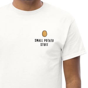 May include: A white short-sleeved t-shirt featuring a small graphic of a brown potato on the upper left chest area. Below the potato graphic, the text SMALL POTATO STUFF is printed in a simple, black, sans-serif font on the white fabric.