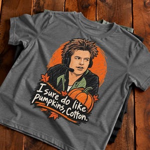 May include: A charcoal gray short-sleeve t-shirt featuring a graphic illustration of a person wearing a headset, set against an orange circular background with a pumpkin and maple leaves. The text reads, "I sure do like pumpkins, Cotton."