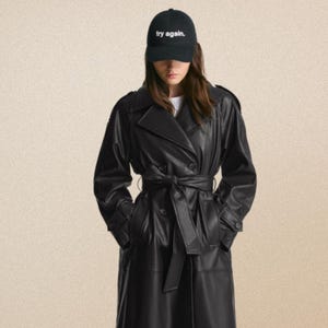 May include: A black trench coat with a matching belt and a black baseball cap. The cap has the text "try again." in white. The coat is made of a shiny material and has a classic trench coat design.