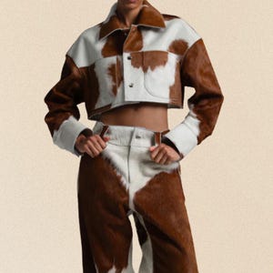 May include: A two-piece outfit featuring a cropped jacket and pants. The jacket and pants are brown and white, with a cowhide pattern. The jacket has a collar and button closures. The pants are high-waisted.