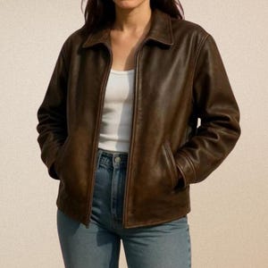Könnte beinhalten: Braune Lederjacke mit Kragen und Reißverschluss. Die Jacke hat zwei Vordertaschen und wird über einem weißen Tanktop und einer blauen Jeans getragen. Die Jacke scheint abgenutzt zu sein.