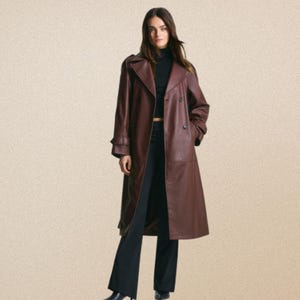 May include: A long, double-breasted, burgundy trench coat. The coat features a classic design with a notched collar, button closures, and a belt. The model is wearing a black turtleneck and black trousers. The coat is knee-length.