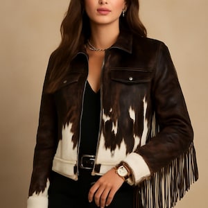 Könnte beinhalten: Braune und weiße Kuhfelljacke mit Fransen. Die Jacke hat einen Kragen, zwei Vordertaschen und einen Reißverschluss. Die Ärmel haben weiße Manschetten, und die Jacke wird mit einem schwarzen Oberteil und einer Hose getragen.