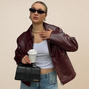 May include: A burgundy leather jacket with a collared neckline and a front zip. The person is wearing a white vest top, blue jeans, black sunglasses, and gold jewellery. A black handbag and a coffee cup are also visible.