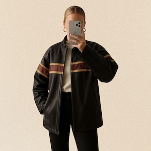 Könnte beinhalten: Schwarze Lederjacke mit braunen und goldenen Streifen auf den Ärmeln. Die Jacke hat einen Reißverschluss vorne und einen Stehkragen. Die Person trägt einen weißen Pullover und eine schwarze Hose.