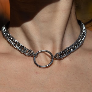 Puede incluir: Un collar gargantilla de cadena plateada con un gran anillo plateado en el centro.