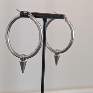 Może przedstawiać: Srebrne kolczyki koła z wisiorkiem w kształcie stożka. Kolczyki są prezentowane na czarnym stojaku na białym tle. Koła są okrągłe i mają polerowane wykończenie.