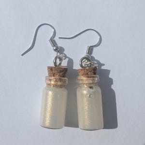 May include: A pair of handmade earrings featuring miniature glass bottles filled with iridescent, pearlescent liquid. Each bottle is topped with a cork and a silver-colored hook for pierced ears. The earrings are designed to dangle.