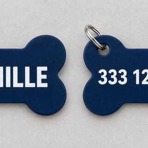 Può includere: Due targhette identificative per animali a forma di osso blu scuro, ciascuna dotata di un anello in metallo argentato. Una targhetta riporta il testo bianco ACHILLE, mentre l'altra mostra la sequenza numerica bianca 333 123 4567 in carattere sans-serif.