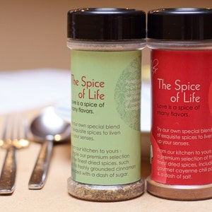 Puede incluir: Dos tarros de especias con tapas negras y etiquetas que dicen "The Spice of Life". El tarro de la izquierda tiene una etiqueta verde y el tarro de la derecha tiene una etiqueta roja. Ambas etiquetas tienen texto sobre las especias que son una mezcla de especias exquisitas para animar tus sentidos.