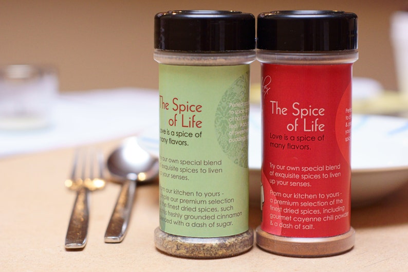 Puede incluir: Dos tarros de especias con tapas negras y etiquetas que dicen "The Spice of Life". El tarro de la izquierda tiene una etiqueta verde y el tarro de la derecha tiene una etiqueta roja. Ambas etiquetas tienen texto sobre las especias que son una mezcla de especias exquisitas para animar tus sentidos.