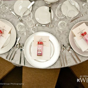 Puede incluir: Una mesa puesta con platos de servicio plateados, platos blancos y servilletas blancas. Cada puesto incluye un peque&ntilde;o frasco de vidrio lleno de caramelos rosas y rojos. La mesa est&aacute; cubierta con un mantel blanco con un patr&oacute;n sutil.