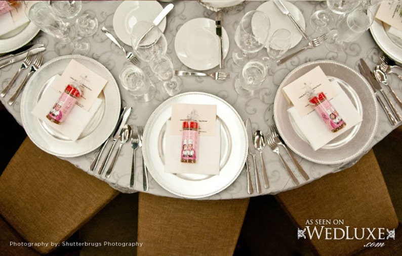 Puede incluir: Una mesa puesta con platos de servicio plateados, platos blancos y servilletas blancas. Cada puesto incluye un peque&ntilde;o frasco de vidrio lleno de caramelos rosas y rojos. La mesa est&aacute; cubierta con un mantel blanco con un patr&oacute;n sutil.