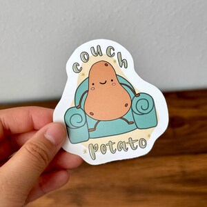 Puede incluir: Una pegatina blanca con una patata de dibujos animados sentada en un sillón azul. La patata tiene una cara feliz y el texto "couch potato" está escrito arriba y abajo de la patata.
