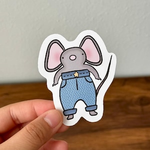 May include: A gray mouse wearing blue denim overalls with a yellow star on the front. The mouse has pink ears and a long tail.